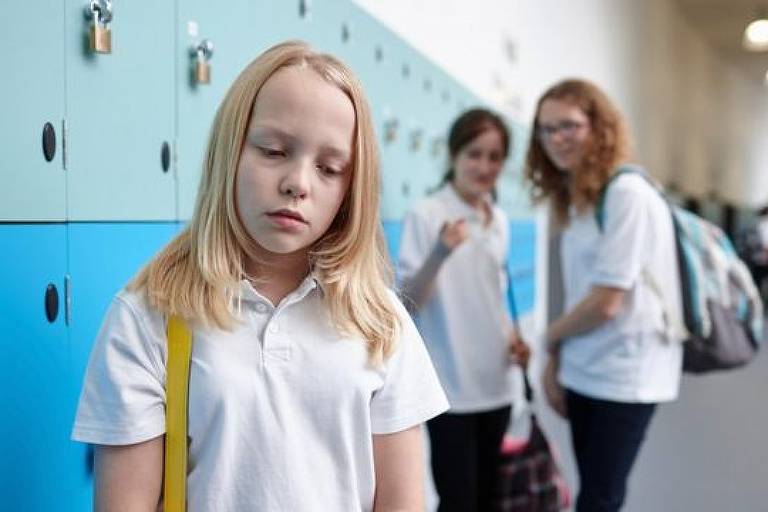 Imagem mostra três adolescentes em um corredor de um colégio. Duas estão rindo e a outra com semblante triste