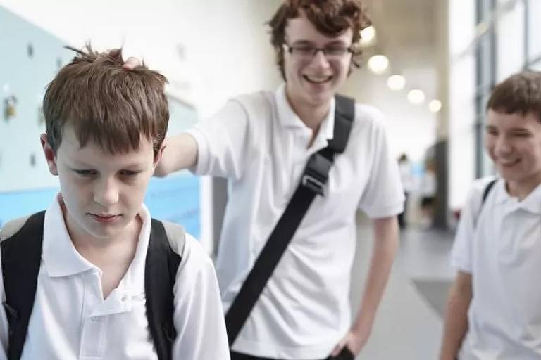 Imagem mostra três adolescentes em um corredor de um colégio. Dois estão rindo e o outro com semblante triste