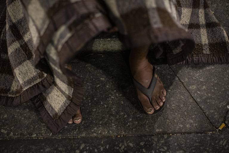 Em uma fotografia feita de noite e de cima, aparece os pés de uma pessoa em pé calçando chinelos pretos e um pedaço de um cobertor com estampa de xadrex.