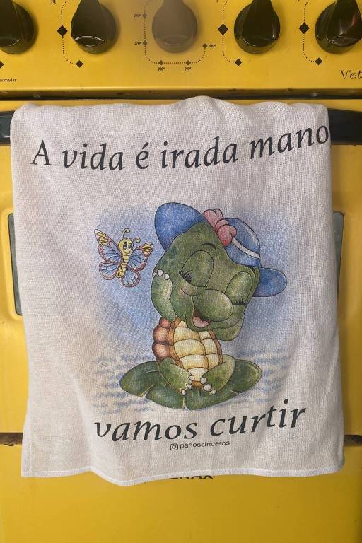 Foto destaca pano de prato pendurado em um fogão; o pano é estampado com o desenho de uma tartaruga e possui os dizeres: 'A vida é irada, mano, vamos curtir'
