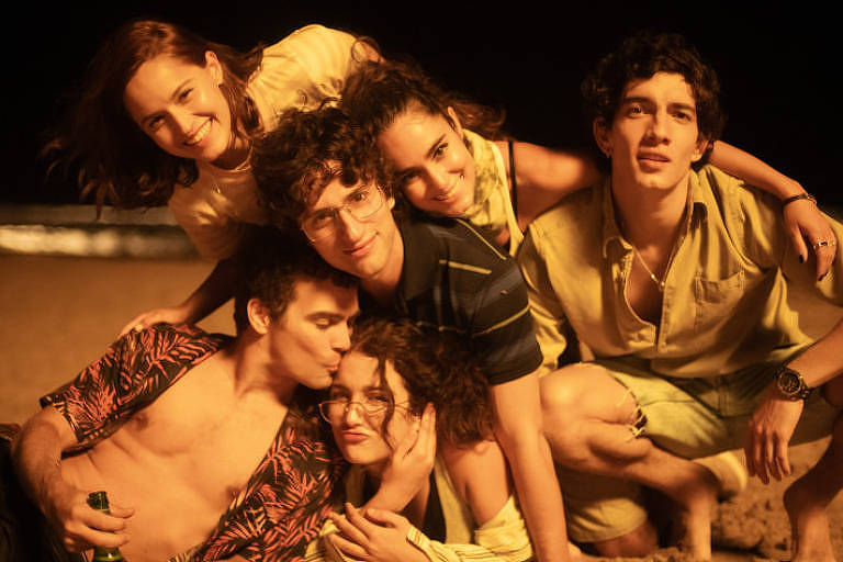 Cena de série representando três homens e três mulheres jovens brancos deitados e se abraçando na praia