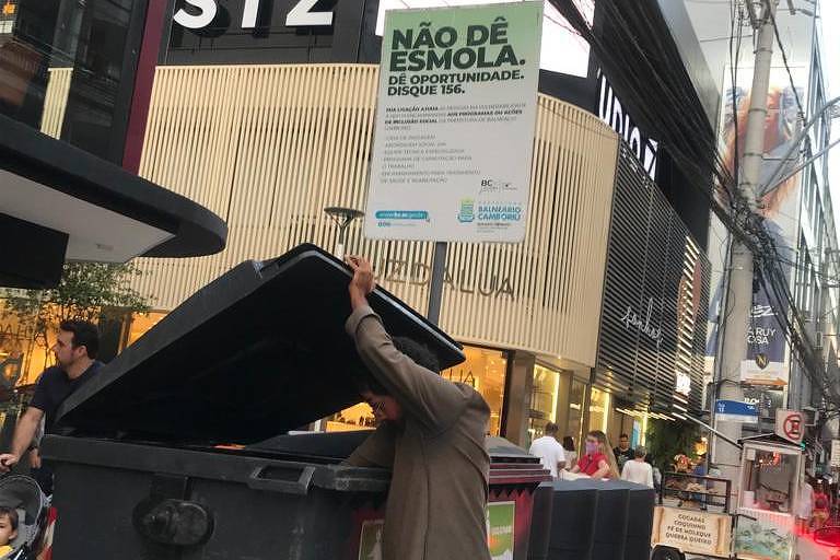 Morador de rua procura coisas no lixo ao lado de placa da prefeitura, em Balneário Camboriú