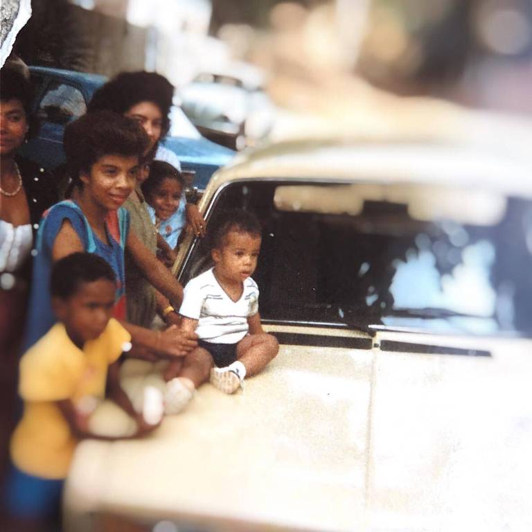 Retrato de neném em cima de capô de carro, com grupo de adultos e crianças ao lado