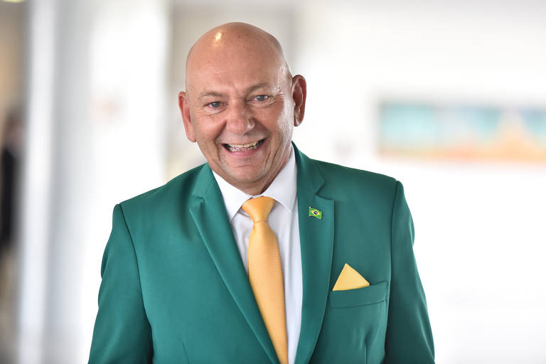 Homem branco, calvo, sorri para a foto. Ele veste um terno verde com gravata e lenço amarelos. A foto é iluminada e o fundo é claro