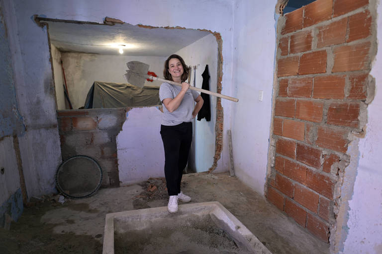 Wanda está no centro de um cômodo em reforma, com paredes sem reboco e cimento concentrado em uma parte do chão; ela segura uma pá usada em obras, que está apoiada sobre os ombros dela