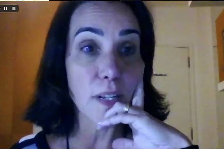 Fotografia colorida de videoconferência mostra mulher em primeira plano; ela é branca, tem olhos e cabelos castanhos escuros, cabelos lisos na altura do ombro; ela tem olhar de supresa e uma das mãos apoiada no rosto