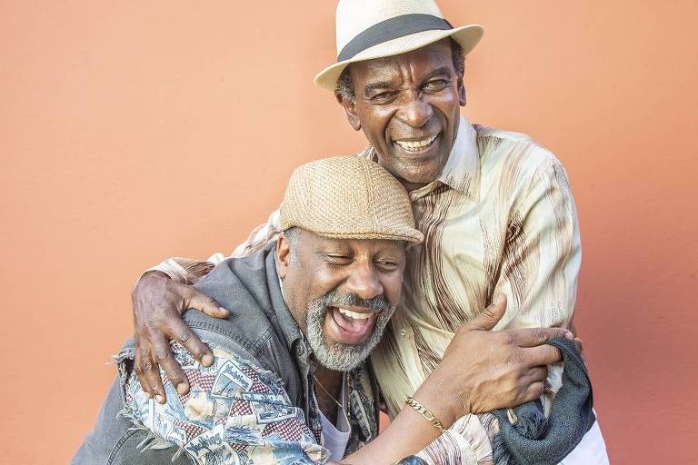 dois homens negros riem e se abraçam diante de fundo laranja