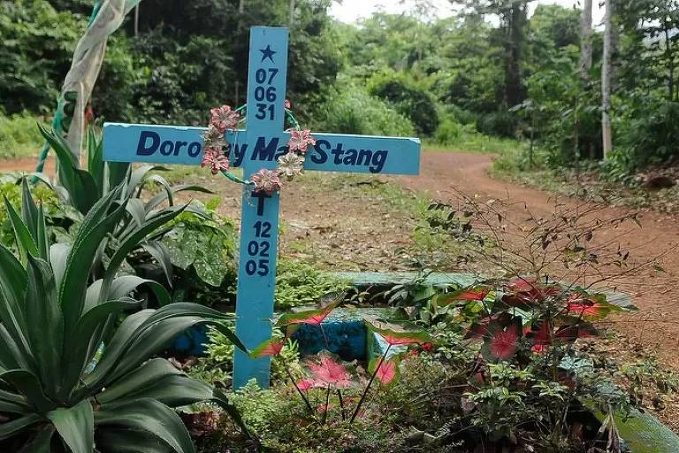 Uma cruz azul com o nome da freira americana Dorothy Stang está no meio de vegetação, ao lado de uma estrada de terra