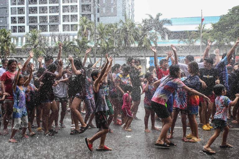 um grupo de mulheres, homens e crianças está em uma rua, recebendo chuva provocada por jatos de água
