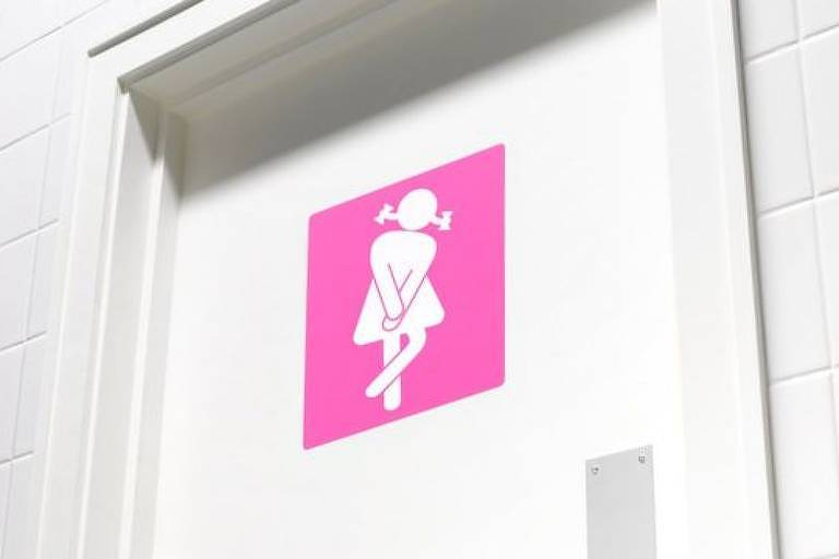 Foto mostra placa quadrada em porta de banheiro com ilustração que representa uma mulher. Ela está em pé, tem as pernas cruzadas e as mãos sobre o ventre.