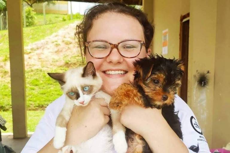 A veterinária usa um jaleco branco e segura um gato e um cachorro nos braços enquanto sorri para a foto