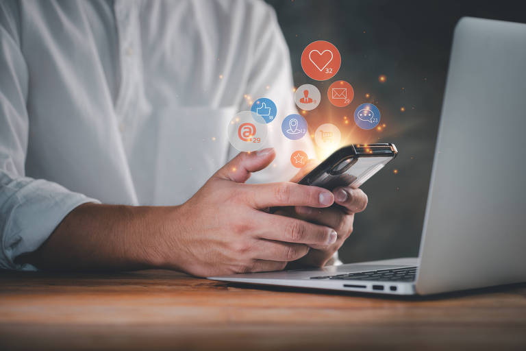 Imagem mostra mão de um homem mexendo em celular; símbolos de redes sociais saem da tela