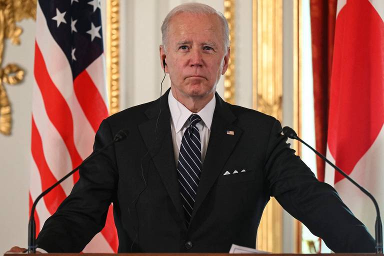 Biden está à frente das bandeiras dos EUA e do Japão; ele veste terno e grava e está próximo de microfones