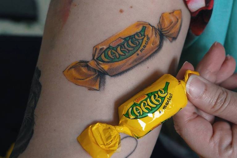 Veterinária Mariane Brasil mostra tatuagem de bombom Caribe em seu braço direito e segura com a mão esquerda o chocolate