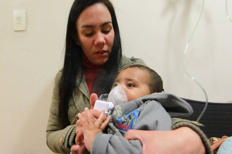 Imagem mostra bebê fazendo nebulização em sala de hospital. Ele está no colo de sua mãe.