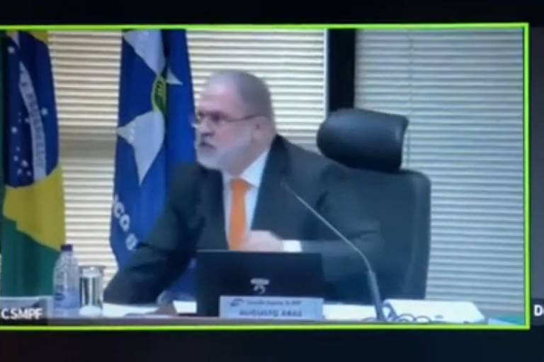 A imagem mostra uma tela de computador em uma videoconferênica; do lado esquerdo está Aras, que veste um terno preto e gravata laranja. Ele está sentando, e faz um movimento brusco para levantar da cadeira; ao lado direito, está um homem careca, com óculos e terno preto. Ele observa a situação