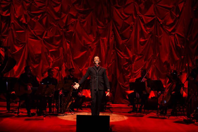 Em foto colorida, o cantor Jean William aparece cantando em um palco iluminado de vermelho