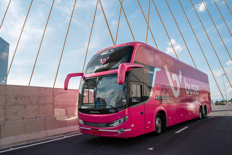 Imagem mostra ônibus de viagem, de dois andares, rosa e com logo da Buser. Ele está sobre uma ponte.