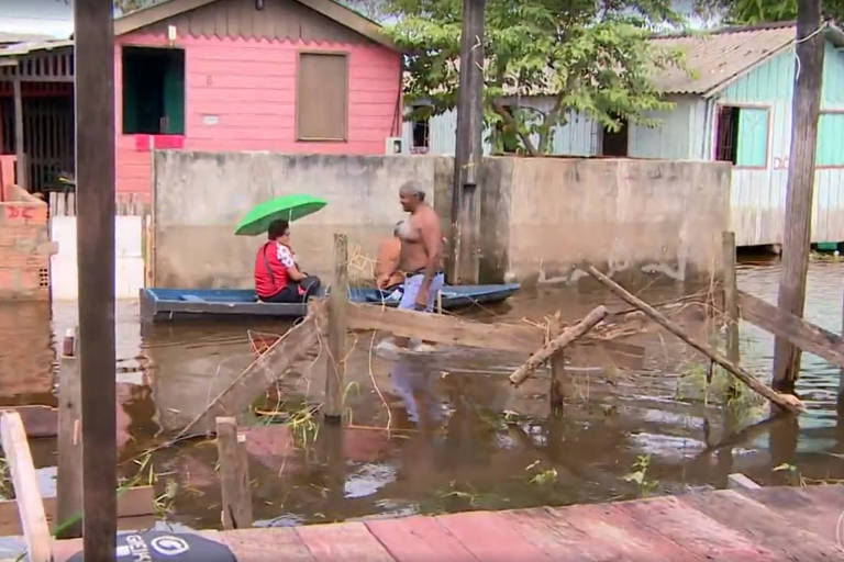 rua alagada com duas pessoas dentro de barco em meio a casas de tijolos
