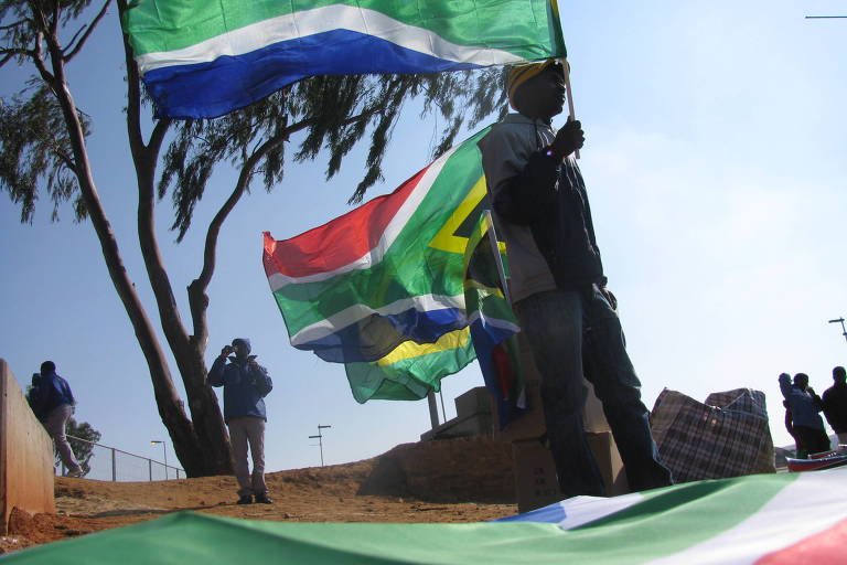 Imagem mostra homem segurando bandeira da África do Sul. há outras bandeiras atrás dele, todas do mesmo país, e uma no chão. Algumas pessoas passeiam pelo lugar.