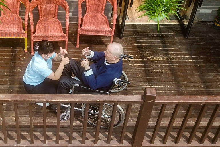 Profissional de saúde fazendo exercícios de reabilitação junto a um idoso, sentado em uma cadeira de rodas; ambos estão em uma varanda