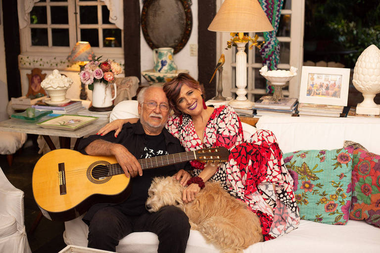 Em foto colorida, o compositor e instrumentista Roberto Menescal e a cantora Cris Delanno posam para a câmera sentados em um sofá com um cachorrinho