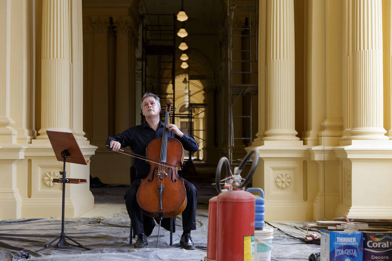 Sobre um chão coberto por um pano preto, rodeado por tintas, um extintor e andaimes, está um homem que, sentado, toca um violoncelo