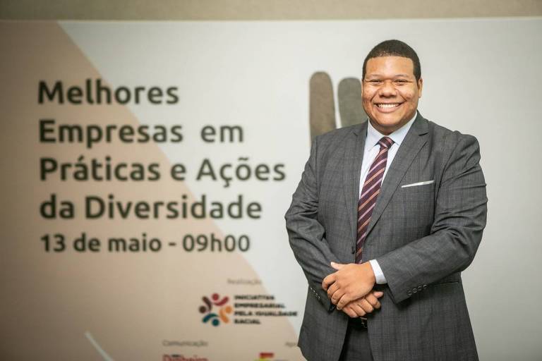 Imagem mostra homem negro, vestido de terno preto e gravata vermelha, com as mãos cruzadas e sorrindo. Ele está em frente a um painel, no qual está escrito "Melhores empresas em práticas e ações da diversidade".