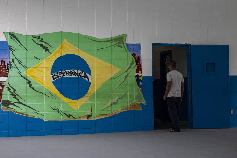 Parece com a bandeira do Brasil com a inscrição "Esperança". Ao lado, uma porta aberta e um jovem vestindo camisa branca de costas