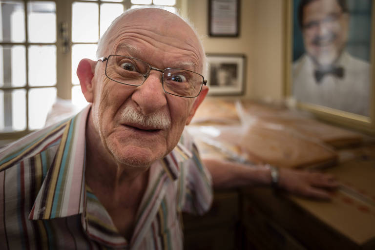 Walter Taverna, um senhor de óculos, olha diretamente para a câmera, enquanto um bolo está na mesa atrás dele