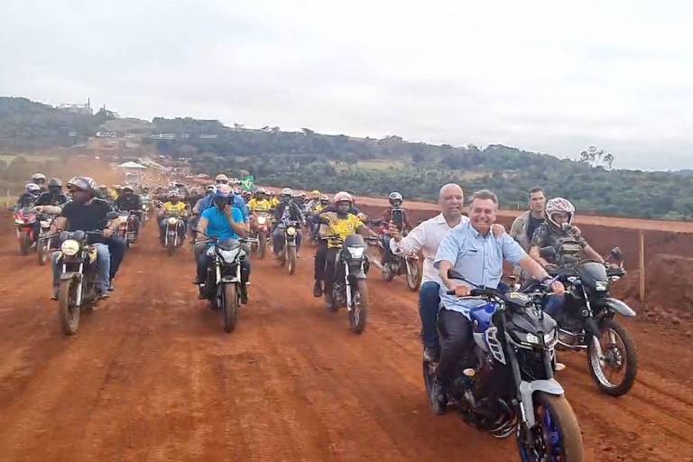 Presidente Jair Bolsonaro (PL), sem capacete, pilota moto em estrada de terra durante visita a Jataí (GO); dezenas de outras motos podem ser vistas atrás dele; há, também, um carona, sem capacete, na moto pilotada pelo presidente