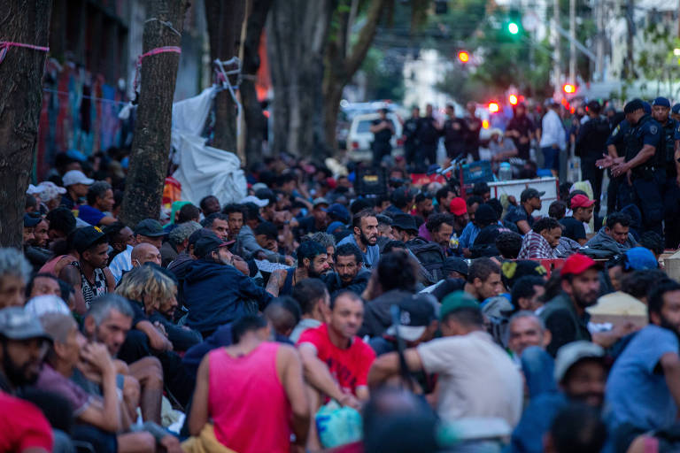 Multidão se concentra na calçada, sentada; ao fundo, guardas civis conversam