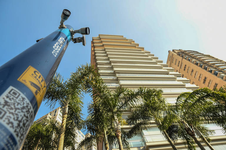 Poste com várias câmeras na ponta, visto de baixo, ao lado de palmeiras e edifício.