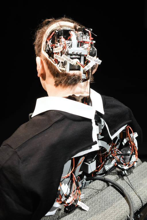 A peça (da foto) é em formato de palestra e conduzida por um humanoide que tem uma aparência bizarramente semelhante à do escritor e dramaturgo alemão Thomas Melle