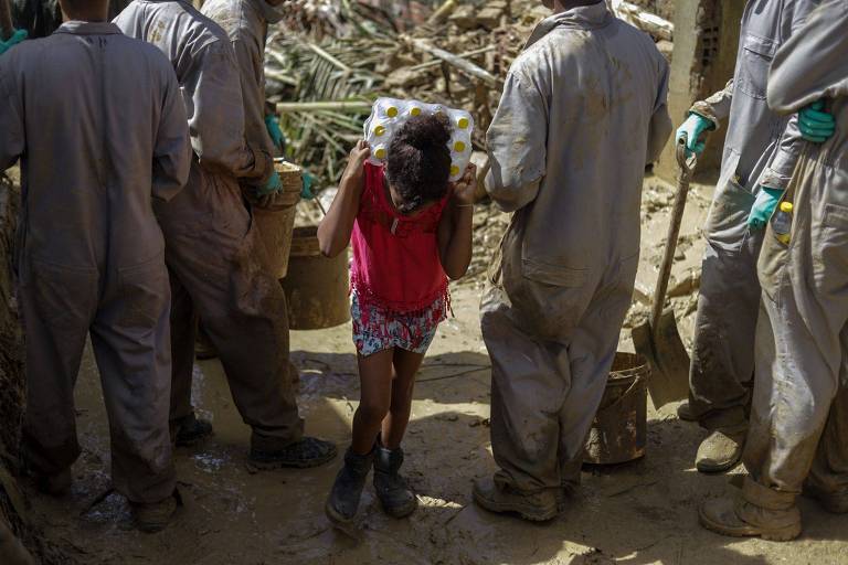 mulher carrega garradas de água entre pessoas com macacão sujo de lama e pás em área de lama