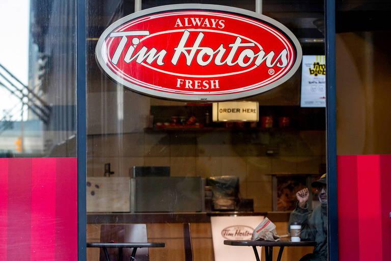 Imagem mostra loja da Tim Hortons. É possível ver janelas de vidro, com um adesivo da logo da marca. Dentro, há cadeiras e mesas de madeira e balcões.