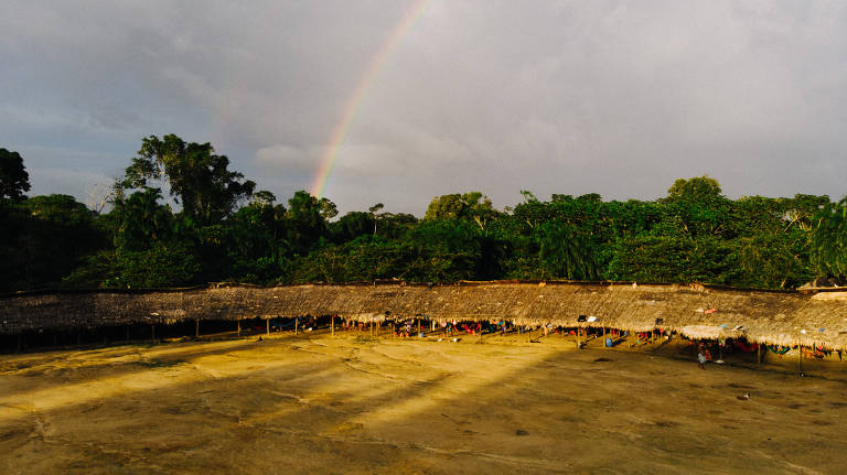 foto mostra terra indígena, com pátio cercado por floresta, e um arco-íris no céu ao fundo