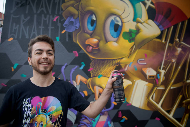 Artista com um jato de tinta em frente a um mural grafitado com o personagem Piu-Piu
