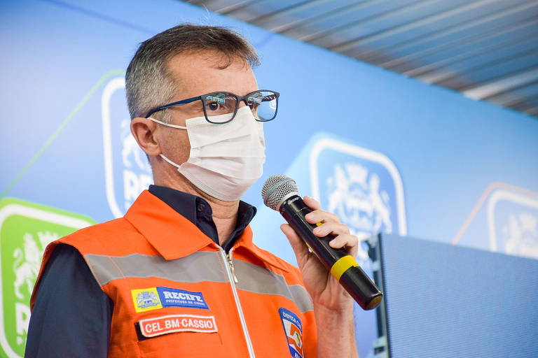 




Cássio Sinomar usando óculos e máscara e com a mão segurando o microfone. Ele usa colete laranja da Defesa Civil sobre a camisa azul