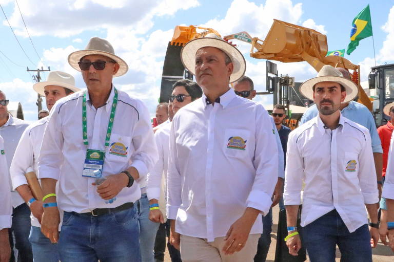 três homens de chapeu e camisa clara com ceu azul e maquina agricola ao fundo, com bandeira do brasil