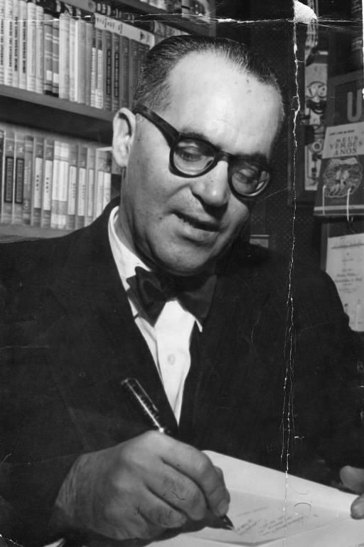 A foto, na vertical, mostra o escritor João Guimarães Rosa em frente a uma estante, enquanto escreve em algum papel. Ele é um homem branco, de cabelos curtos e cabeça já calva, que usa óculos e terno com gravata borboleta.