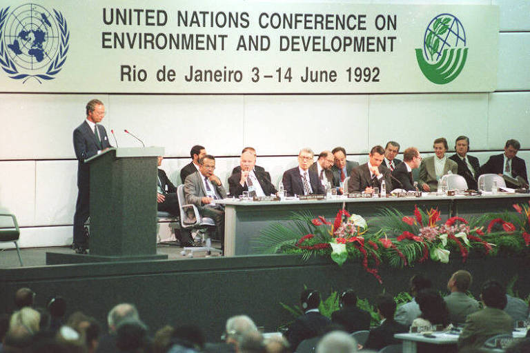 Em um palco um homem discursa na tribuna enquanto outros estão sentados diante de uma longa mesa; ao fundo há um painel com o nome da conferência, em inglês