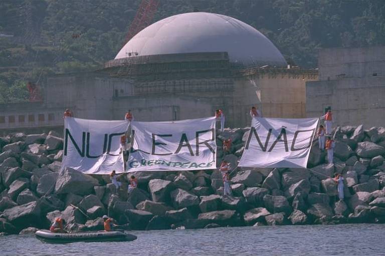 Panos brancos com a frase 'Nuclear Não', escrita em preto, são segurados por pessoas de pé em pedras diante do mar; ao fundo é possível ver o edifício da usina