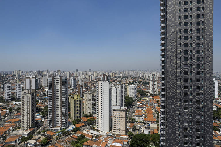 Imagem mostra paisagem cheia de prédios sob um céu azul. No canto direito da foto, é possível ver um prédio em destaque, maior que os outros. Ele é cinza e corta toda a foto verticalmente.