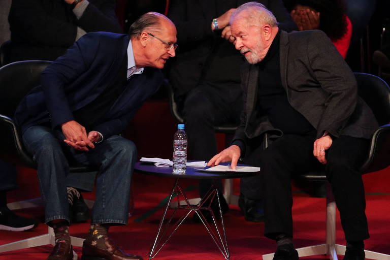 Alckmin, à esquerda, e, Lula, à direita; os dois estão sentados e se aproximando para conversar
