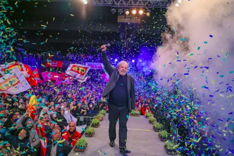 Lula em passarela, vem em direção à câmera com mão direita levantada; ao redor da passarela, multidão com bandeiras e papéis picados voando