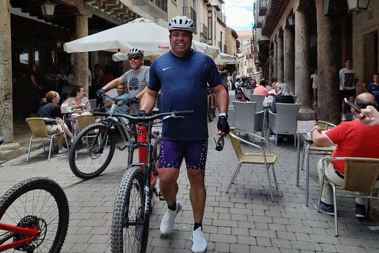 O ex-jogador Ronaldo aparece em pé ao lado de uma bicicleta elétrica vermelha de última geração; ele usa bermuda e camisa justas, como os ciclistas profissionais, e usa capacete protetor na cabeça 