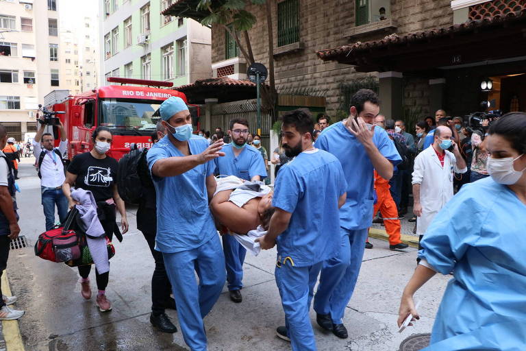 Funcionários de uniformes azuis de hospital carregam homem em uma maca; ao fundo, está o caminhão dos Bombeiros