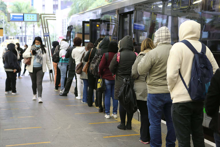 fila de passageiros na diagonal, de costas, da direita para a esquerda, ao lado deles há um ônibus