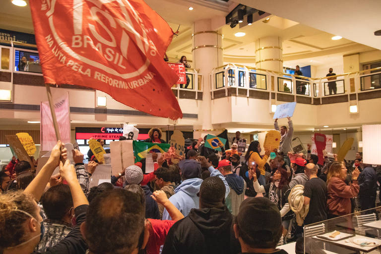 Imagem mostra protestantes do MTST dentro do shopping Iguatemi. Eles estão reunidos com cartazes, e uma bandeira vermelha com o logo do movimento está hasteada. 